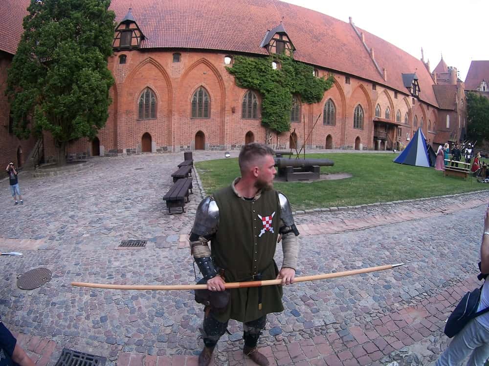 Dziedziniec zamku średniego. Rycerz drzewcem włóczni toruje przejazd.  W tyle mury zamku.