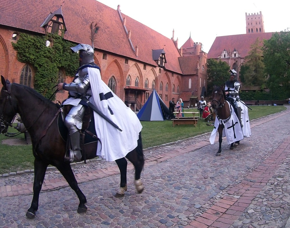 Rycerze zakonu na koniach przemierzają dziedziniec zamku średniego
