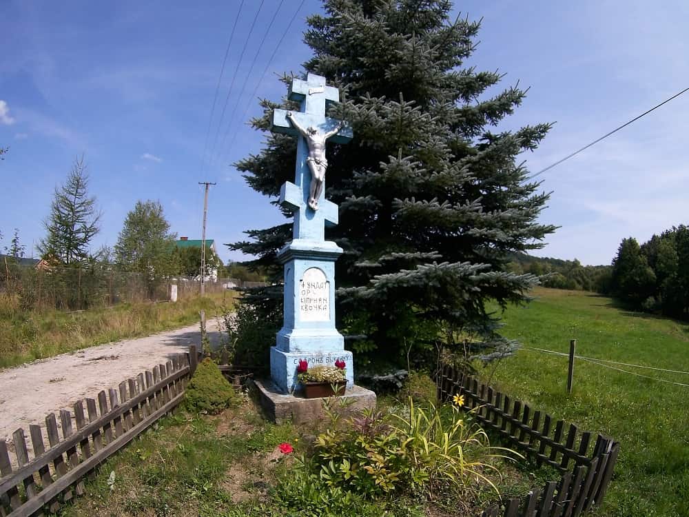 Krzyż o ośmiu końcach i trzech poprzeczkach wykonany z kamienia. na krzyżu postać Jezusa. Krzyż otoczony jest drewnianym płotkiem.
