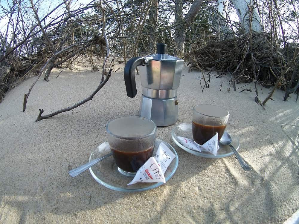 Kawiarka i dwie filiżanki z mocną kawą. Wszystko to stoi na piasku. w tle suche gałązie.