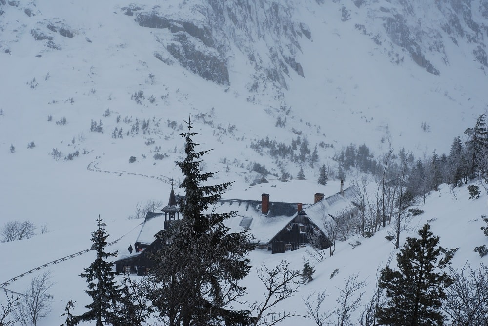 Widok schroniska PTTK Samotnia od strony szlaku z góry. Wdać charakterystyczną wieżyczkę. Na pierwszym planie choinki w tle zarys Małego Stawu skutego lodem.