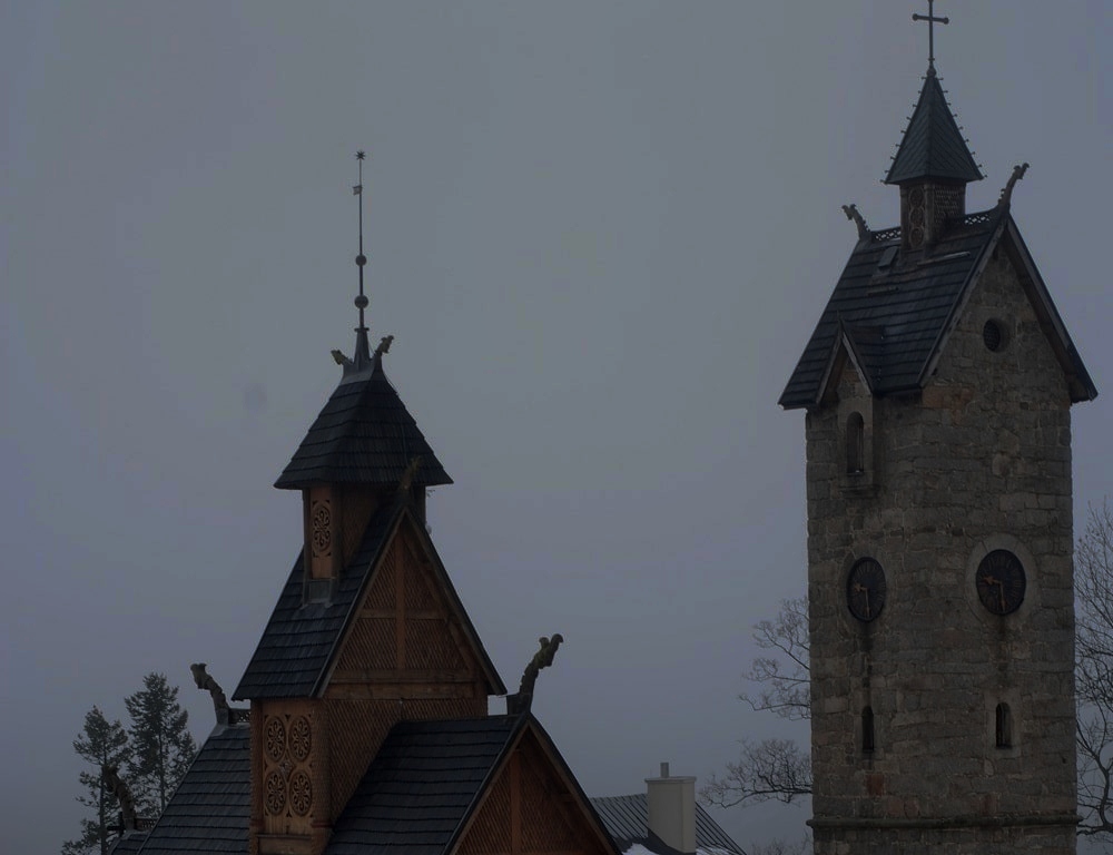 Po lewej kamienna wieża z zegarem wskazującym 21.30, która chroni świątynię przed kaprysami Ducha Gór. Po lewej, jakby na dalszym planie, drewniana świątynia z charakterystycznymi zdobieniami. wszystko pogrążone w mroku/