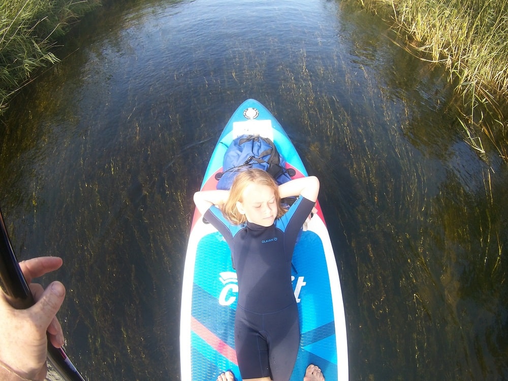 Zuzanna leży na płynącej desce SUP. płynie z ojcem. Jego nie widać na fotografii, może za wyjątkiem dłoni w lewym dolnym rogu. Deskę otacza woda, to właśnie rz. Piaśnica 
fot. Rafał Lipkowski TJNB