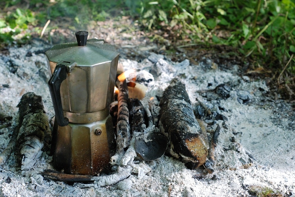 Kawiarka na żarze wśród niemal zwęglonych patyków. Widać lekki płomień i zieleń poza niemal białym popiołem.