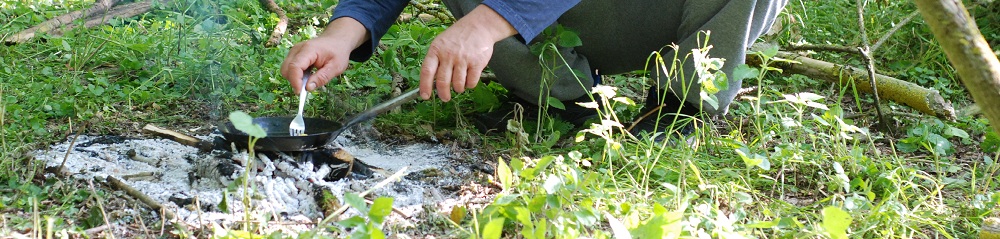 Patelnia na żarze ogniska. Jedna dłoń przytrzymuje patelnię, druga pracuje widelcem. nie widać postaci jednak widać jak przyrządza jajecznicę